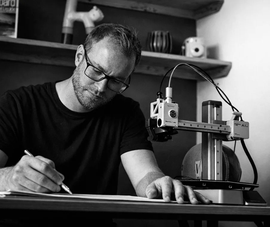 Jonny sketching at his desk beside a small desktop 3D printer. Black and white photograph.